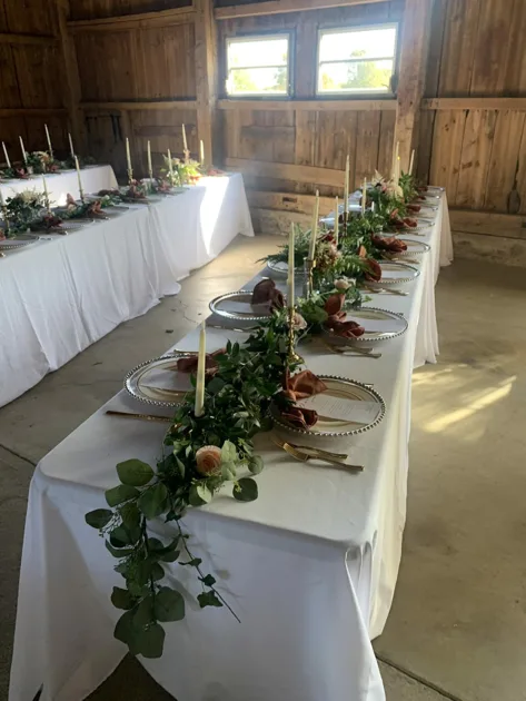 Green Mixed Leaf Garland White Tablecloth White Candlesticks Roes Clear Plates Beaded Rim Charger Plates Side Angle View Greenery Centerpiece